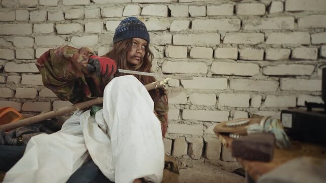 castaway child in dirty white trousers and camo jacket sharpens thick wood with large cutlass while sitting near brick wall, beside scattered kitchen items and pot on burner inside cold unkempt