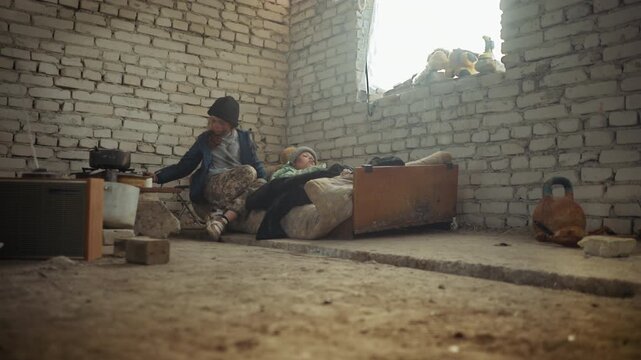 Older girl gives water to sick sister lying on mattress near window in cold brick room while another person in background holds cutlass near makeshift stove surrounded by worn cooking tools