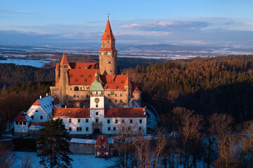 Bouzov Castle aerial winter view at golden hour, medieval fortress with red roofs above forest landscape, Czech Republic.