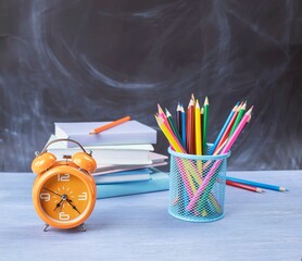 Back to school concept, stack of books, colored pencils and alarm clock on table near blackboard.