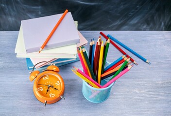 Back to school concept, stack of books, colored pencils and alarm clock on table near blackboard.