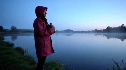 Standing by lake holding thermos. Woman in raincoat with hood at dawn. Mist and reflection soften light. Image evokes wellness and outdoor lifestyle. Suitable for wellness travel lifestyle brand use.