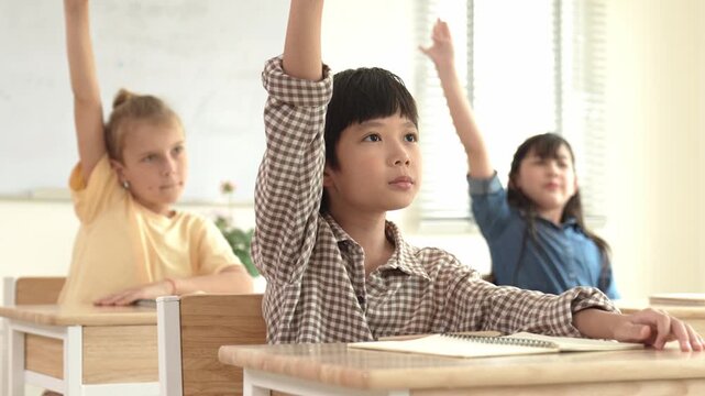 Smart boy raising hand for asking and answering teacher surrounded by multicultural children studying at classroom. Happy student working together, voting, volunteering, calling instructor. Pedagogy.
