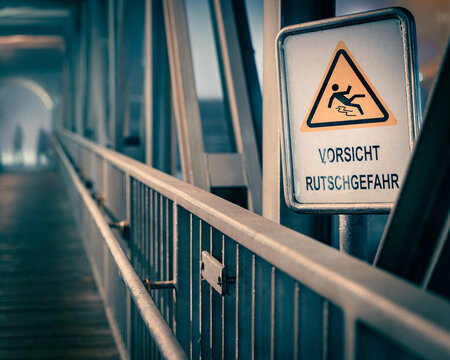 Warnschild &bdquo;Vorsicht Rutschgefahr&ldquo; auf einer Br&uuml;cke/einem Steg, starke Tiefenunsch&auml;rfe, urbane industrielle Umgebung, horizontal