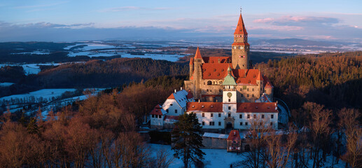 Bouzov Castle aerial winter view at golden hour, medieval fortress with red roofs above forest landscape, Czech Republic.
