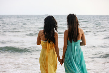Two woman in summer dresses holding hands stand on a beach on sea waves background