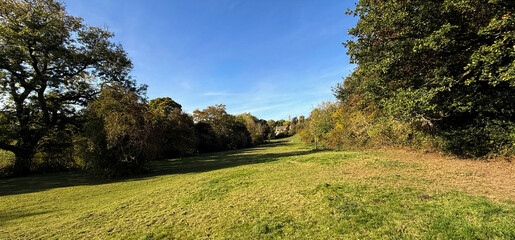 A wide grassy pathway stretches far into the distance, flanked by lush green trees. The autumn sky is a clear blue, enhancing the vibrant colors in Norwood Green, Halifax, UK