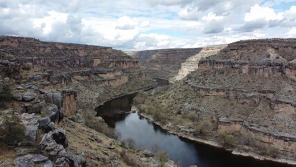 High-angle panoramic view of a dark river winding through a deep rocky canyon under a cloudy sky. The landscape features steep, rugged limestone or sandstone cliffs with sparse vegetation, capturing t