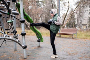 A man in a tracksuit and hat stretches with boxing gloves on an outdoor sports field. Exercising in cold weather.