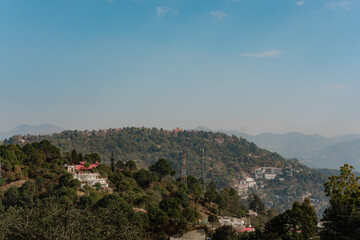 Fototapeta premium Scenic Mountain Landscape with Lush Greenery and a Clear Blue Sky, Serene Vista of Rolling Hills and Scattered Buildings, Perfect for Travel and Nature