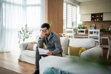 Fototapeta premium Man feeling overwhelmed reading financial papers at home