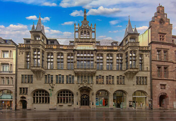 Obraz premium Historic architectural building in Basel with symmetrical facade, ornate details, and reflections on a quiet city street under a cloudy blue sky.