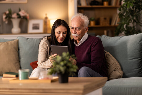 Adult daughter explaining to her senior father how tablet is working while they sitting on couch together in living room.