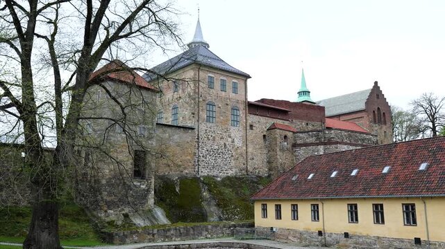 layered historic stone fortress buildings rises above mossy rock foundations red tiled roofs yellow barracks church spires norwegian flag appear akershus oslo view norway 