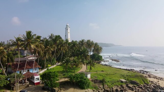 Aerial drone photo of Dondra Head Lighthouse in Sri Lanka with traditional fishing boats and fishermen in the foreground. Tall white lighthouse standing on the southernmost point of the island.