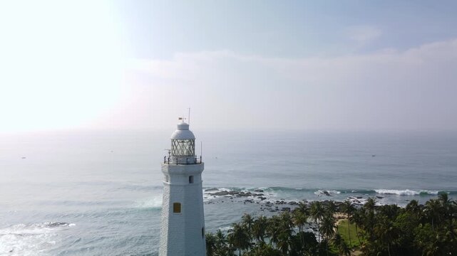 Aerial drone photo of Dondra Head Lighthouse in Sri Lanka with traditional fishing boats and fishermen in the foreground. Tall white lighthouse standing on the southernmost point of the island.
