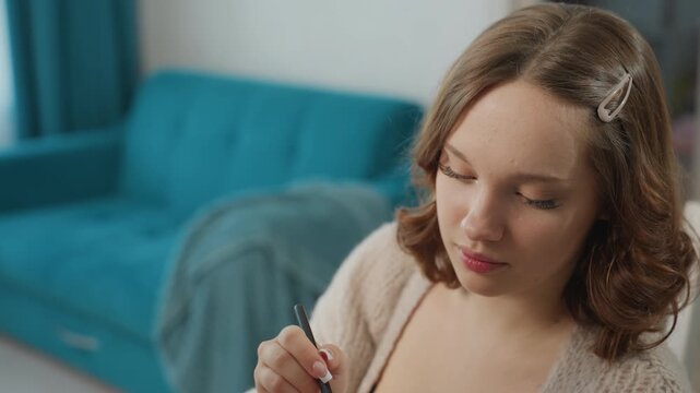 Caucasian Woman Holding Eyeliner Pencil, Sitting On Teal Sofa, Concentrating On Precise Eye Line, Soft Window Light, Hair Clip, Cozy Sweater, Closeup Facial Detail, Home Beauty Routine And Calm Mood.