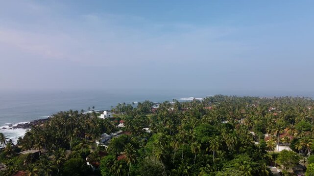Aerial drone photo of Dondra Head Lighthouse in Sri Lanka with traditional fishing boats and fishermen in the foreground. Tall white lighthouse standing on the southernmost point of the island.