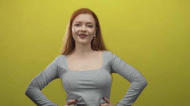 Redhead woman posing with hands on hips showing bare upper chest and square neckline in a studio with a yellow green backdrop and slight smile; confidence.
