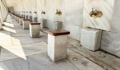Row of brass water taps with stone seats arranged for ritual ablution. Religion, culture, and daily...