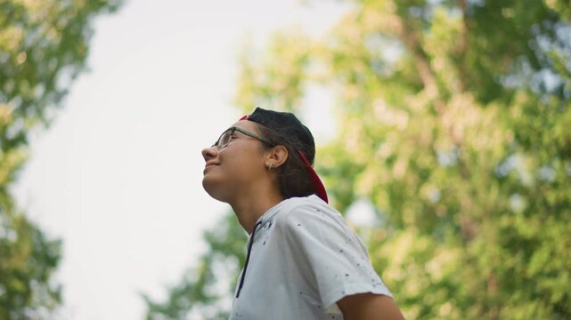 young person looking up at trees, wearing glasses and backward cap, soft smile, sunlight through canopy, backpack strap on shoulder, breath of fresh air, hopeful contemplative vibe