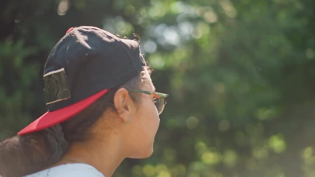 young person wearing cap in forest, student daydreaming and sketching, soft bokeh sunlight, warm rimlight on profile, hooded casual outfit, gentle breeze through greenery, candid side views