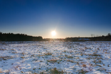 Łąka pokrywa warstwą śniegu spod kt&oacute;rego miejscami wystają suche trawy. W oddali widać las. Nisko nad horyzontem widać zachodzące słońce.