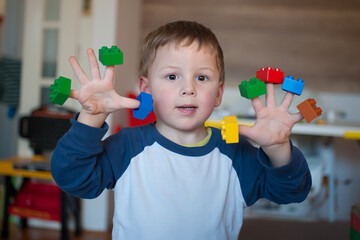 Naklejka premium A young boy holds various small building blocks on his fingers in a room filled with toys. He smiles while playing and enjoying his time with colorful toys