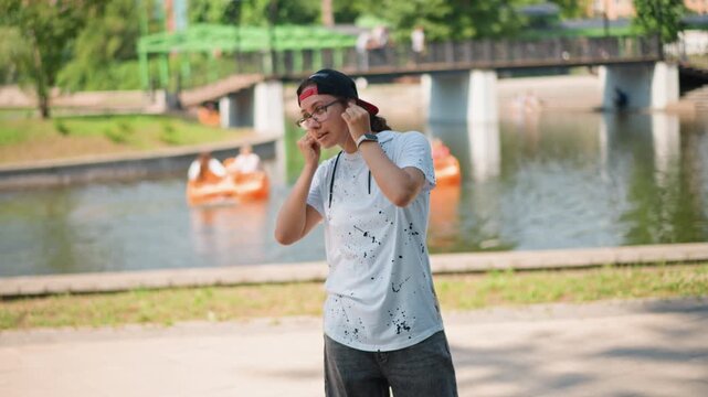 asian man relaxed by riverside boats and bridge, adjusting sunglasses and stretching, casual tee, leisurely stroll, paddleboats in background, calm reflective mood, summer park leisure portrait