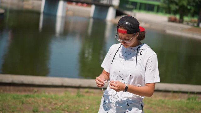 young woman untangling earbuds by pond, adjusting cap and red headband while checking cords and audio before recording sunny urban park, calm water reflection, relaxed posture, lifestyle broll