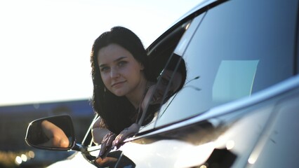 Young beautiful girl leaning out of driver window at modern electric car and looking into camera. Portrait of attractive smiling woman in alternative electrical vehicle. Slow motion