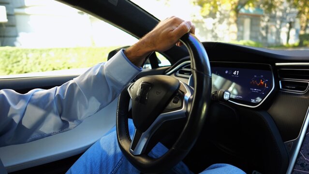 Male hand of businessman holds steering wheel while driving electrical vehicle at city. Businessperson operating electric car on urban road. Young man commuting to work at sunny day. Close up