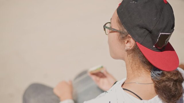 young woman with skateboard sitting outdoors wearing backward red cap and glasses, curly hair in low bun, holding phone on lap, skateboard deck beside, overhead shallow focus, warm beige tones,