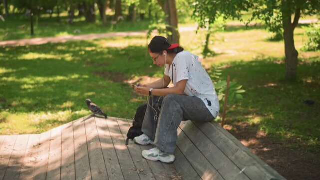 Male youth enjoying peaceful outdoor moment, Teenage male pauses on park bench with device in hand, Young adult sitting on wooden seat reviewing messages beneath leafy trees and bright sunlight