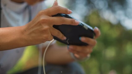 closeup hands holding mobile controller outdoors, focused player using earbuds and cable, sunlight filtering through trees, shallow depth of field, thumb pressing buttons with tense calm - Powered by Adobe
