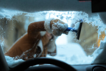 A gloved hand uses a snow brush to clear the windshield, seen from behind the steering wheel, ensuring visibility for winter driving.