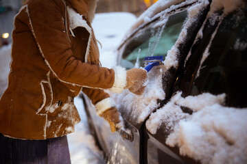Close-up of a person in warm mittens and a suede jacket using an ice scraper on a car window during a cold winter evening or early morning.