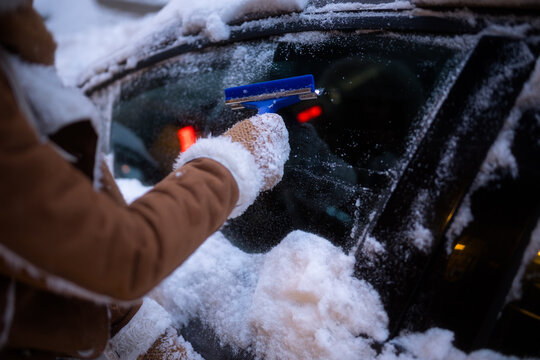 A person in warm clothing uses a blue tool to scrape ice and snow off a car windshield at night, with city lights blurred in the background.