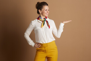 A smiling middle-aged woman, professionally dressed in a white shirt and mustard yellow trousers with a colorful scarf, confidently presents or offers with an open palm.