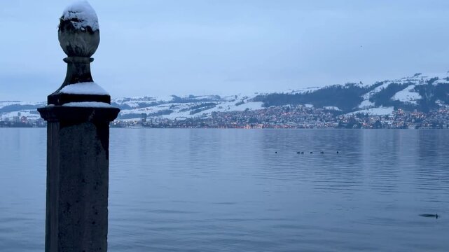 Icy winter conditions at the shore of Lake Zug near H&uuml;nenberg, with snow-covered ground, a calm lake, and the Zugerberg emerging through mist in a mystical atmosphere.
