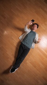 Vertical video young man relaxing on a wooden parquet floor, holding a mobile phone and taking a self portrait, capturing a moment of social media engagement or casual downtime