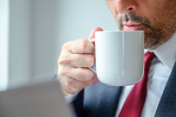 Professional man in suit drinking coffee from mug by window, focus on cup in elegant office setting.