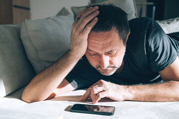 Man lying face down on sofa, hand on head, staring blankly at smartphone, concept for depression and burnout or mobile phone addiction doomscrolling.