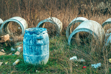 Abandoned blue plastic barrel and concrete pipes in overgrown meadow