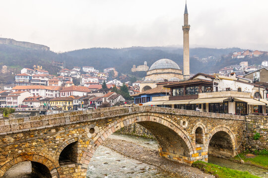 Stone arch bridge crossing a shallow Bistrica river with Ottoman Sinan Pasha mosque and tall minaret rising above dense hillside housing and traditional architecture on overcast day, Prizren, Kosovo