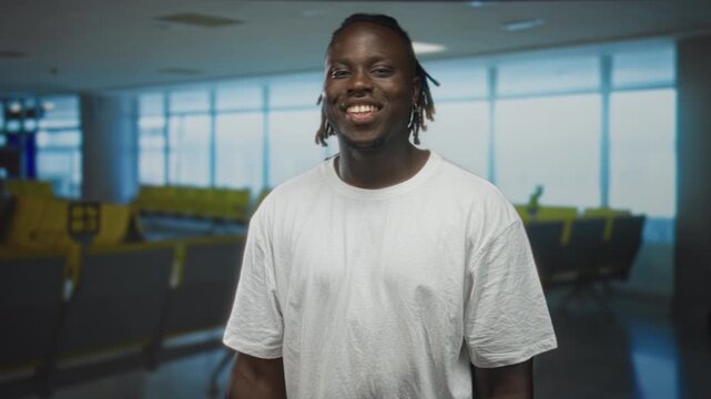 Man with pucker mouth looking up in airport terminal wearing white tshirt and dreadlocks, standing near seats and windows; surprise curiosity.