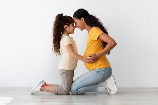 Excited little daughter touching mom's pregnant belly, bonding with mother at home, happy young middle eastern family awaiting for second child, posing near white wall background, side view