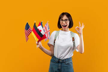 Emotional lady showing bunch of diverse flags and okay gesture, posing with backpack over yellow studio background, happy female student recommending foreign language studying classes