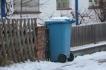 blue plastic recycling bin for paper waste stands at the roadside, covered in snow, ready for collection