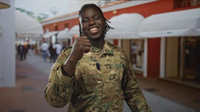 Young man soldier in camouflage uniform gives thumbs up on a busy street lined with white awnings and shops; pride service.